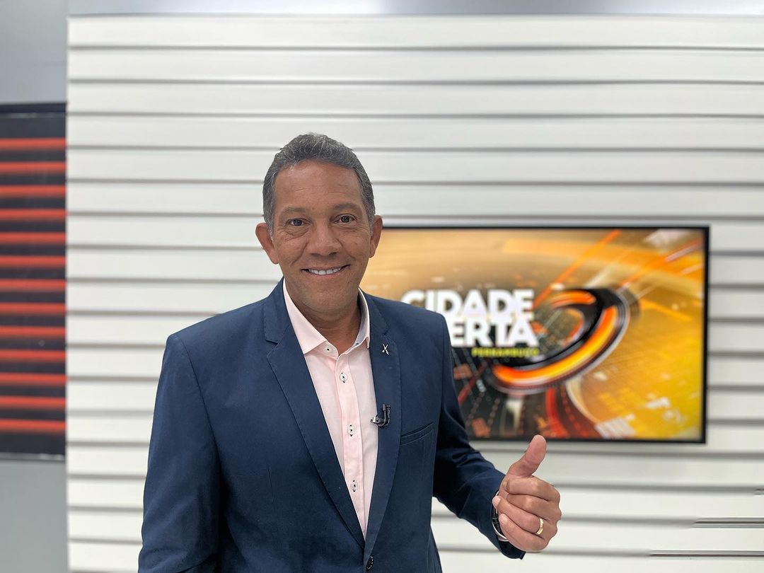 Washington Gurgel no cenário do Cidade Alerta PE. Ele está sorrindo e apontando para a tela onde aparece o logo do telejornal