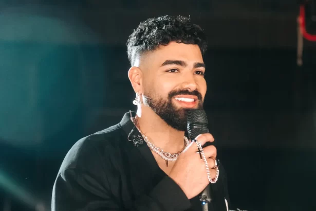 Dilsinho de jaqueta e blazer preto, segurando o microfone, sorrindo, durante ensaio fotográfico