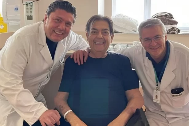 Faustão de camisa azul, sentado numa cadeira de roda, sorrindo, ao lado de médicos em um hospital