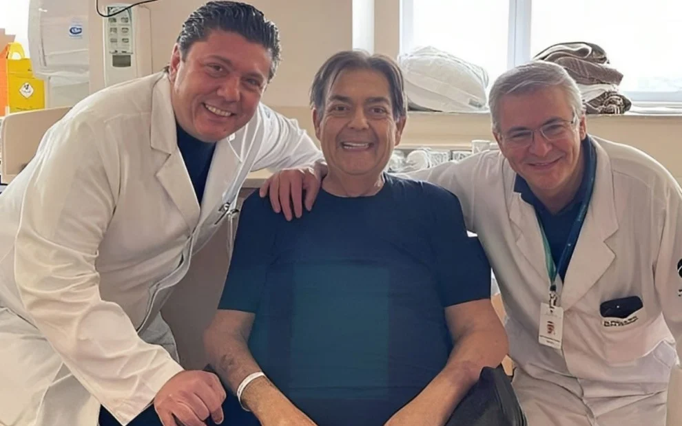 Faustão de camisa azul, sentado numa cadeira de roda, sorrindo, ao lado de médicos em um hospital