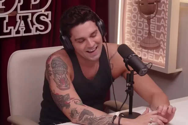 Luan Santana sorrindo, sentado proximo de um microfone, de camisa regata preta, durante entrevista para o PodDelas