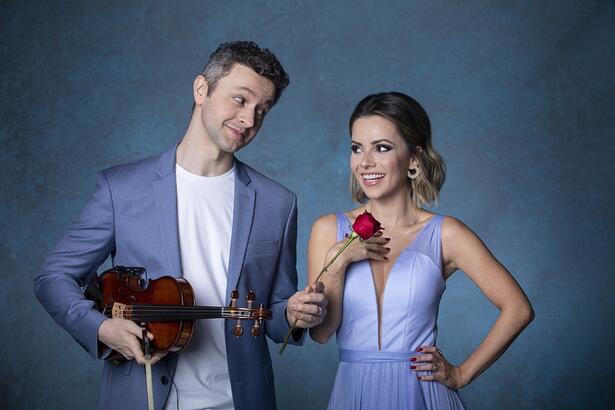 Sandy de vestido azul, segurando uma rosa com a mão direita ao lado de Lucas Lima, de blazer e calça azul e camisa branca, segurando um violino