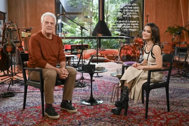 Pedro Bial de camisa marrom, calça bege, sorrindo, sentado ao lado de Sandy, de vestido branco com detalhes pretos, sorrindo, durante gravação do Som Brasil