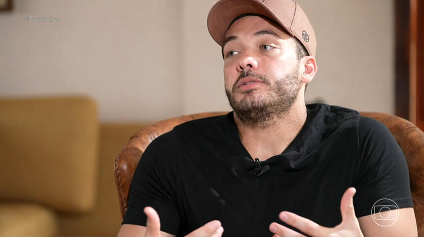 Wesley Safadão de boné marrom, camisa preta, sentado num sofá marrom, durante gravação de entrevista para o Fantástico, da TV Globo