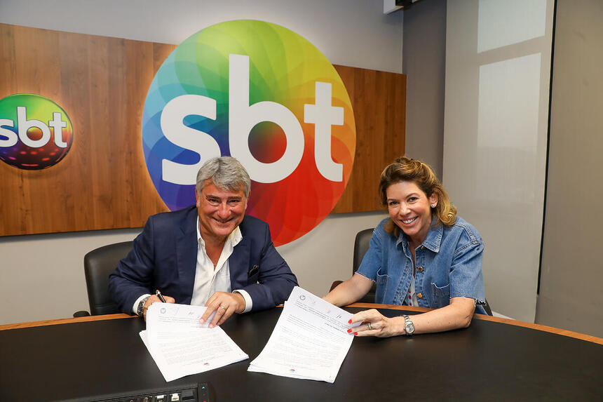 Cleber Machado na sala de Daniela Beyruti, assinando contrato com o SBT. Os dois estão sorridentes sentados à mesa no local que tem o logo da emissora