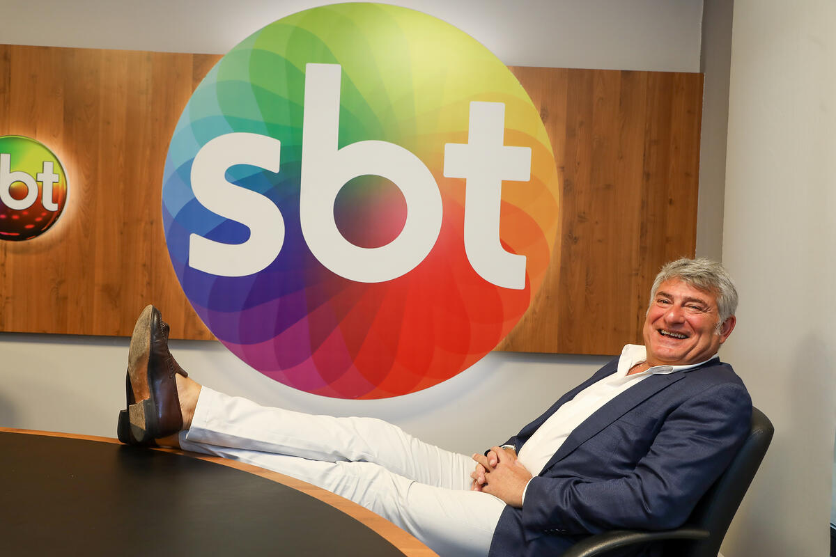 Cleber Machado sorrindo com os pés em cima da mesa do SBT, após assinar com o canal