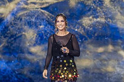 Ivete Sangalo sorrindo, com um vestido preto brilhoso no centro do palco do The Masked Singer Brasil