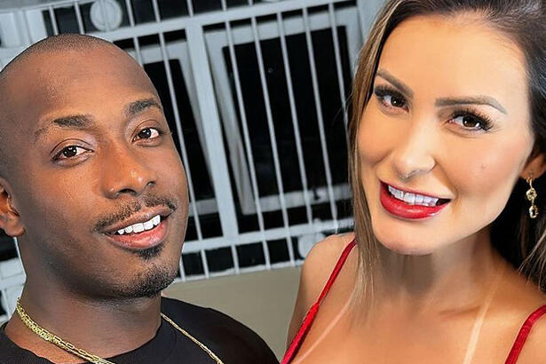 Andressa Urach de camisa regata vermelha, sorrindo, com cabelo liso e solto ao lado de Jeão, sorrindo, de camisa preta ao lado de Andressa