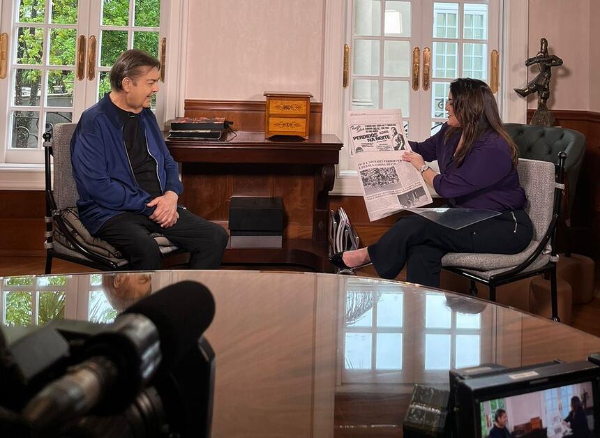 Faustão sorrindo enquanto Fabíola Reipert lendo um jornal antigo durante gravação de entrevista para o Domingo Espetacular