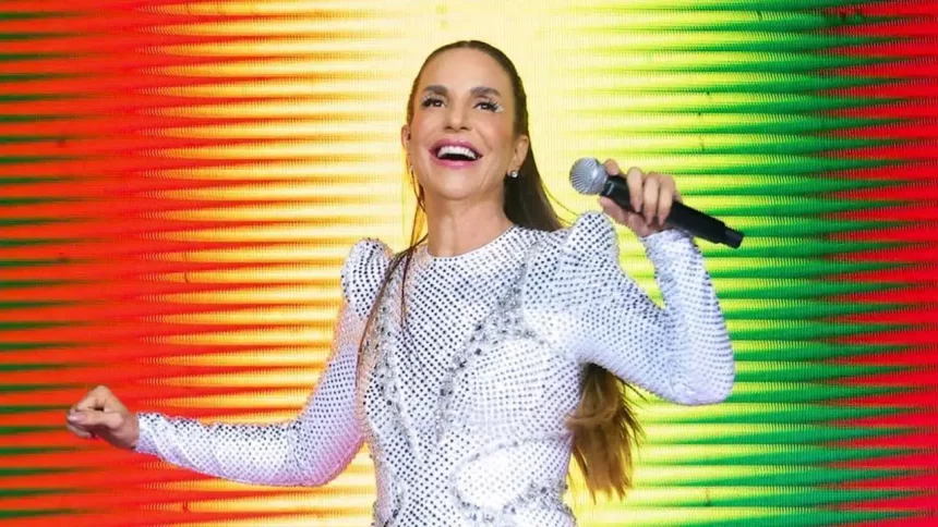 Ivete Sangalo de look branco, com os braços abertos, segurando um microfone, durante show