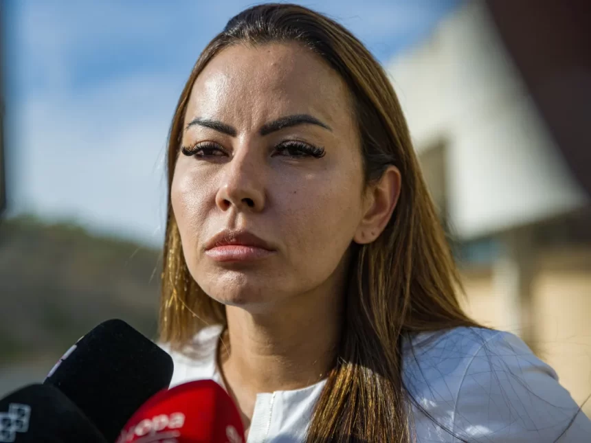 Dinorah Santana de camisa branca, com semblante sério, cabelo liso, solto, dando entrevistas em frente a vários microfones