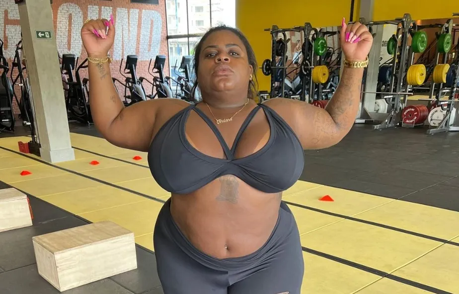 Jojo Todynho com os braços abertos, usando look de academia na cor cinza, em pé, na academia, com semblante sério