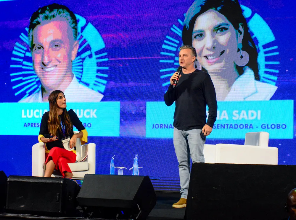 Luciano Huck em pé, de camisa preta manga longa, calça jeans, no palco da Rio Innovation Week