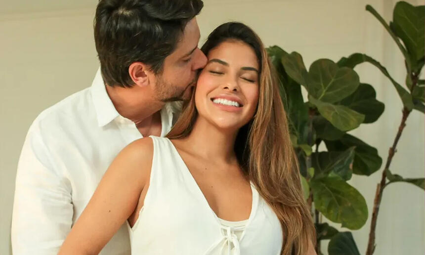 Munik sorrindo, de vestido branco, sendo abraçada pelo marido Paulinho Simões