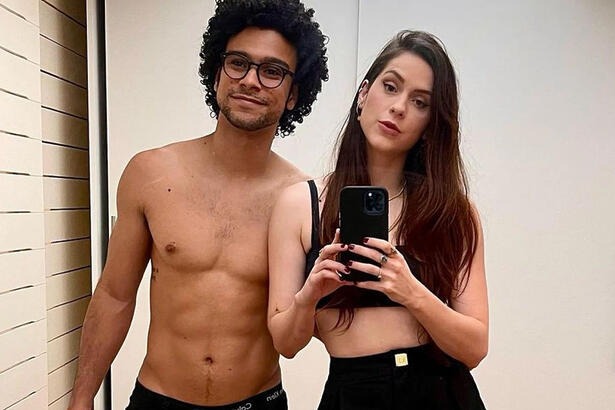 Sophia Abrahão segurando o celular com as duas mãos, usando um top e calça pretos, ao lado de Sério Malheiros, sem camisa, de calça preta, usando óculos de sol