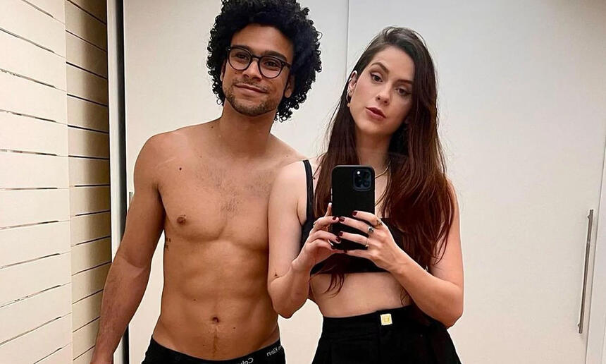 Sophia Abrahão segurando o celular com as duas mãos, usando um top e calça pretos, ao lado de Sério Malheiros, sem camisa, de calça preta, usando óculos de sol