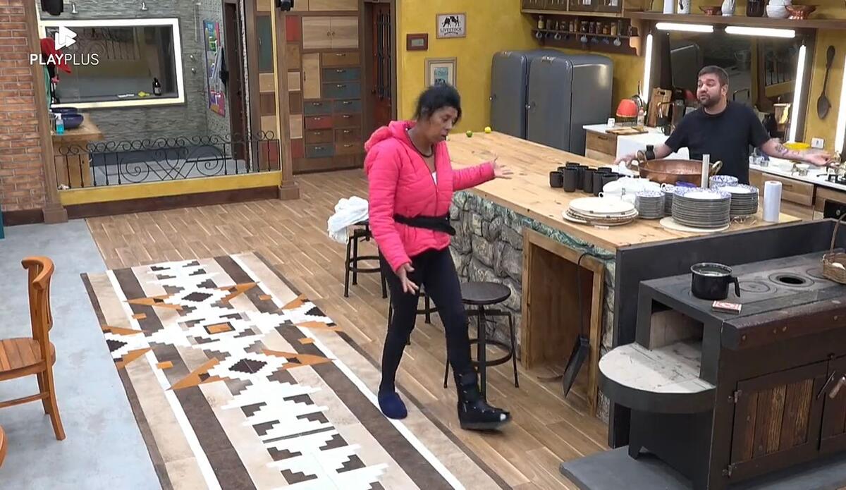 Marcia Fu de uma lado da cozinha em Pé, com uma blusa rosa escura, apontado o dedo para Sander, que se encontra do outro lado perto do fogão.