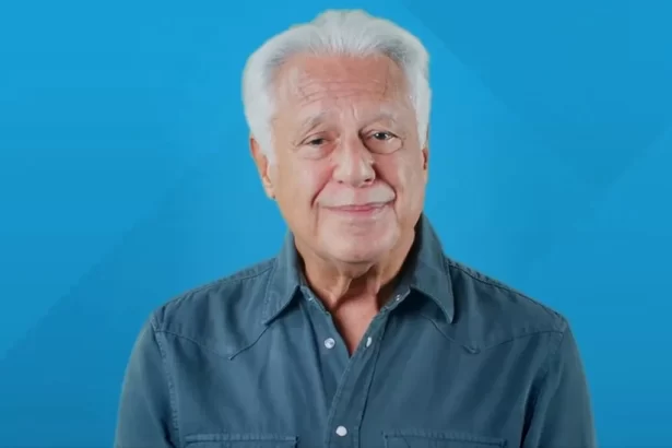 Antonio Fagundes de cabelo branco, camisa social azul/cinza, em estudio com plano de fundo azul, com semblante duvidoso durante gravação de campanha