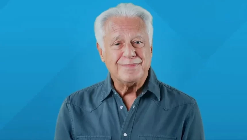 Antonio Fagundes de cabelo branco, camisa social azul/cinza, em estudio com plano de fundo azul, com semblante duvidoso durante gravação de campanha