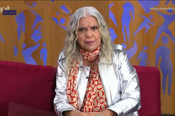 Astrid Fontenelle com o cabelo solto, blazer prateado sentelante, sentada num sofá com pano vermelho no estudio do GNT enquanto grava o Saia Justa