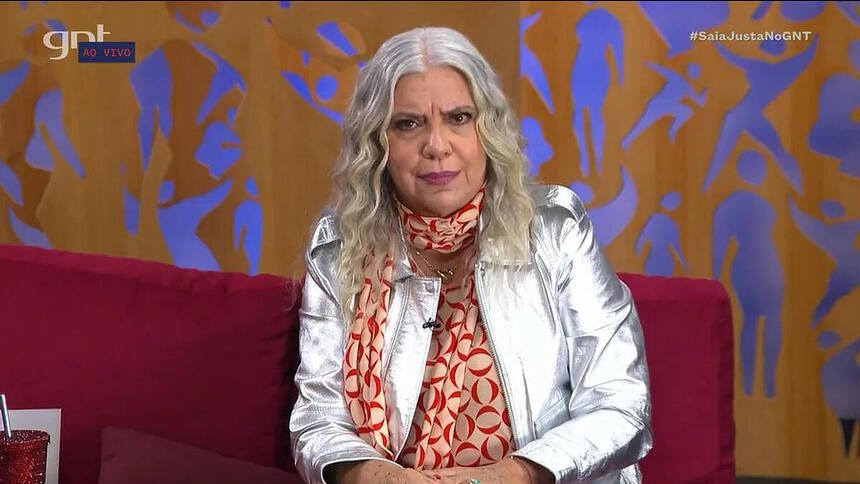 Astrid Fontenelle com o cabelo solto, blazer prateado sentelante, sentada num sofá com pano vermelho no estudio do GNT enquanto grava o Saia Justa