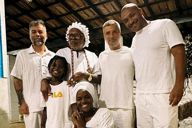 Bruno Gagliasso sorrindo, com roupa toda branca acompanhado de amigos, assessor e filho num terreiro de candomblé