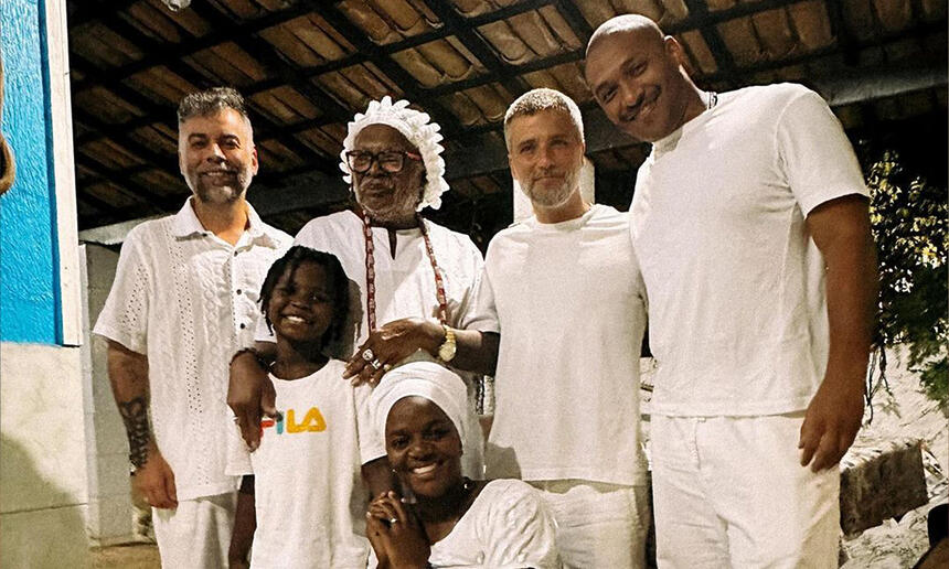 Bruno Gagliasso sorrindo, com roupa toda branca acompanhado de amigos, assessor e filho num terreiro de candomblé