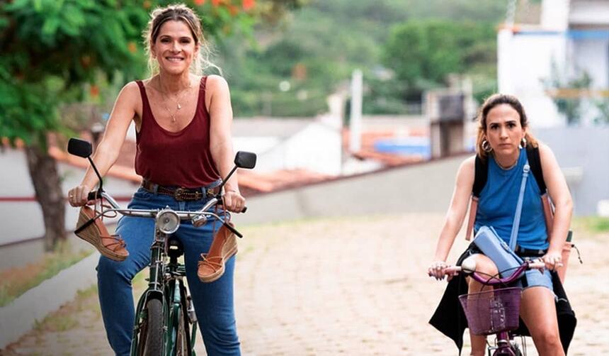 Ingrid Guimarães sorrindo, de camisa regata vermelha, calça jeans, segurando numa bicicleta parada numa praça; Imagem 2: Tatá Werneck de camisa regata, jaqueta jeans amarrada, encostada na bicicleta