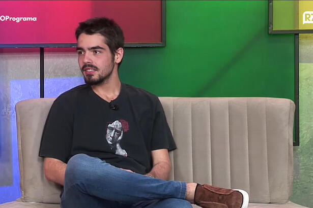 João Guilherme Silva de camisa preta, pernas cruzadas, sentado durante entrevista em podcast