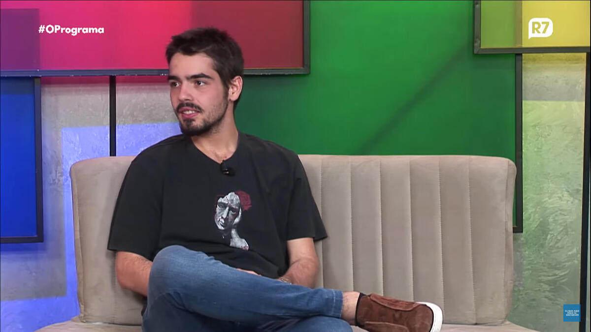 João Guilherme Silva de camisa preta, pernas cruzadas, sentado durante entrevista em podcast