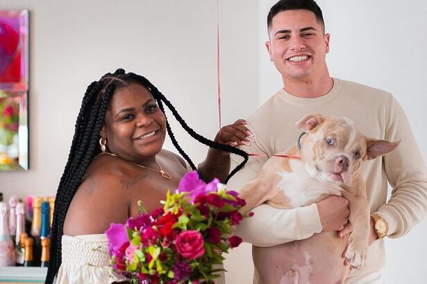 Jojo Todunho com penteado de trança, sorrindo, segurando um buquê de flores coloridas, ao lado de Lucas Souza, que está segurando um cachorro no braço