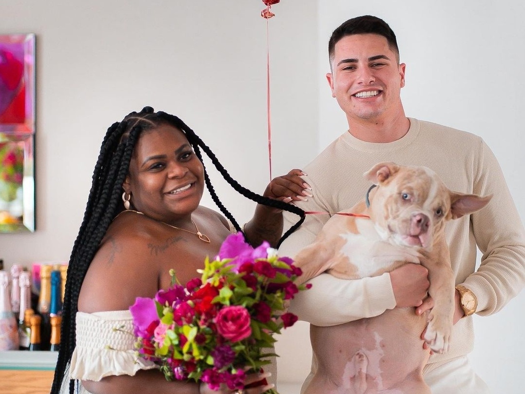 Jojo Todunho com penteado de trança, sorrindo, segurando um buquê de flores coloridas, ao lado de Lucas Souza, que está segurando um cachorro no braço