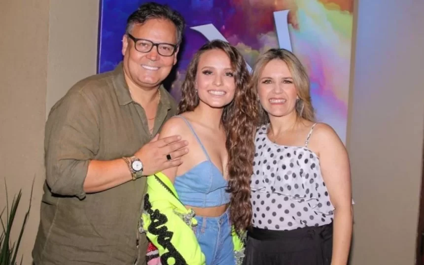 Larissa Manoela sorrindo, de cabelo solto, com o casaco aberto, ao lado dos pais