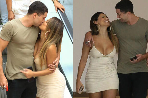 Lexa de vestido curto, bege, de cabelo solto, descendo a escada rolante abraçada com Ricardo Viana