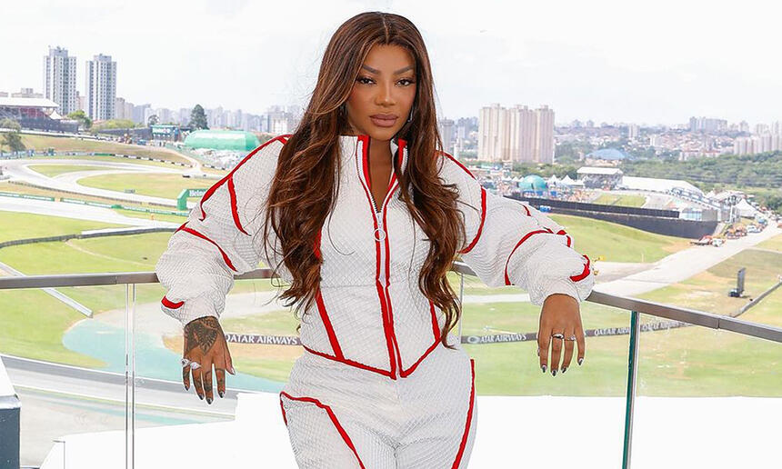 Ludmilla de look branco com detalhes vermelhos, cabelo solto, com semblante sério, encostada num ferro nos bastidores da GP SP 2024