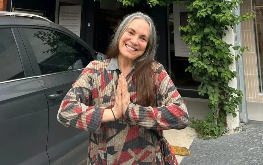 Regina Duarte sorrindo, com as mãos juntas, de camisa estampada em cores marrom, verde e magente