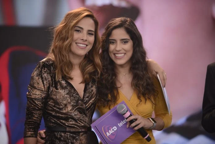 Wanessa de vestido preto com transparencias, sorrindo, de cabelo solto, ruivo, abraçada com Camilla Camargo durante gravação