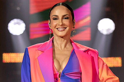 Claudia Leitte de blazer laraja, roxo e rosa, com decote por baixo, sorrindo, segurando um microfone durante gravação do The Voice Brasil, da TV Globo