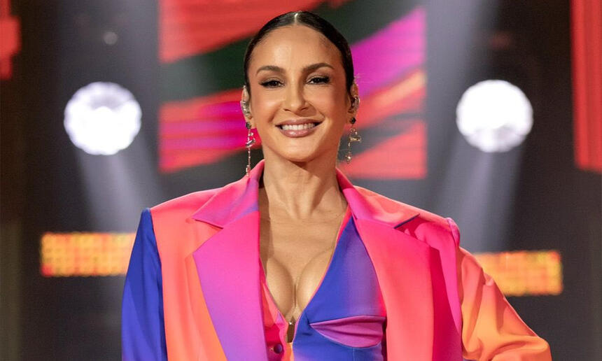 Claudia Leitte de blazer laraja, roxo e rosa, com decote por baixo, sorrindo, segurando um microfone durante gravação do The Voice Brasil, da TV Globo