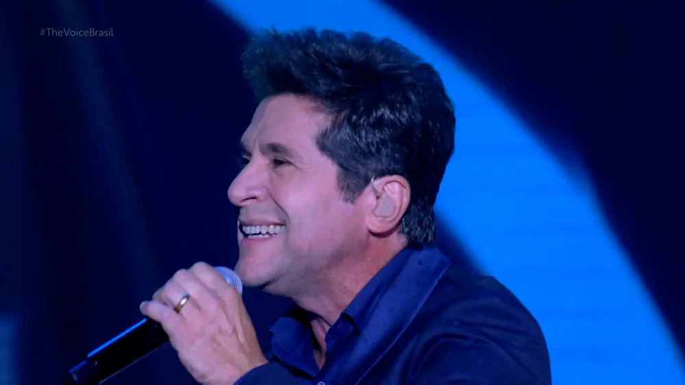 Daniel de blazer azul, de perfil, segurando microfone durante performance no The Voice Brasil