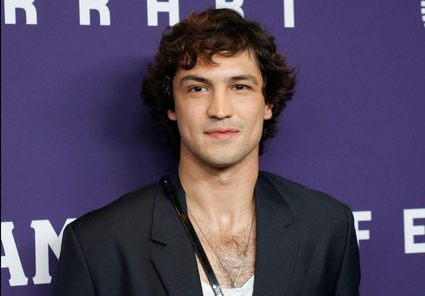 Gabriel Leone de camisa branca e blazer preto em sobreposição, cabelo preto, mais cheio, com semblante feliz em coletiva de imprensa