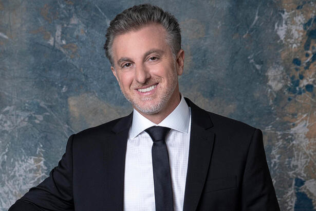 Luciano Huck de blazer e gravata pretos, sorrindo, durante ensaio fotográfico par a emissora