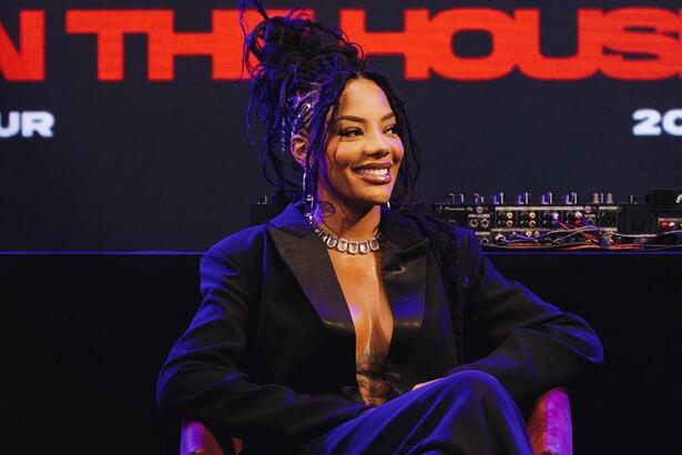 Ludmilla usando trança, de blazer decotado preto, calça social preto, sentada, com as pernas cruzadas, sorrindo, durante coletiva de imprensa da nova turnê