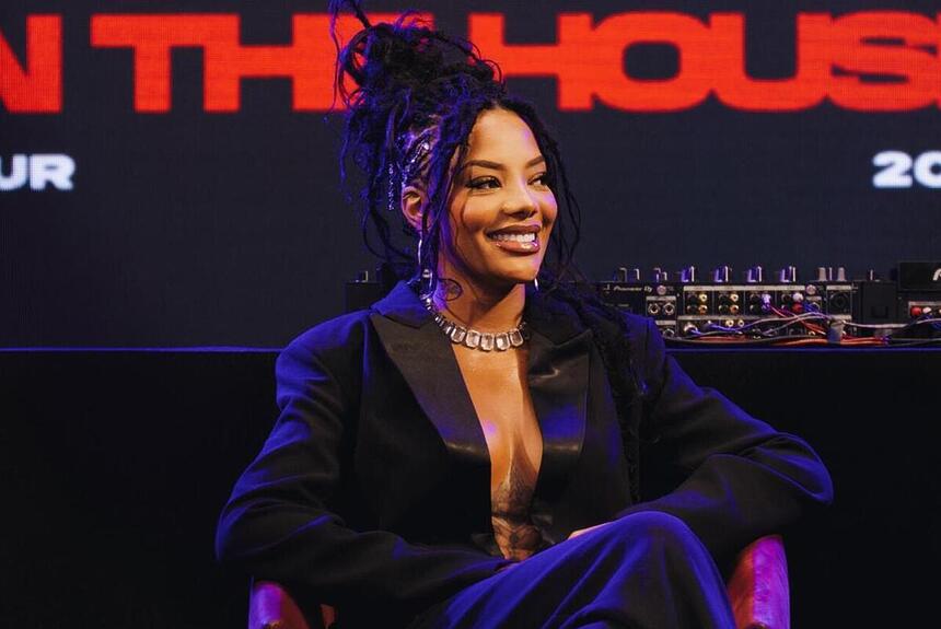 Ludmilla usando trança, de blazer decotado preto, calça social preto, sentada, com as pernas cruzadas, sorrindo, durante coletiva de imprensa da nova turnê