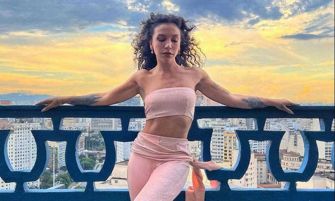 Priscilla Alcantara de top e calça rosa bebê, com os braços abertos, olhos fechados e cabelo solto, durante viagem