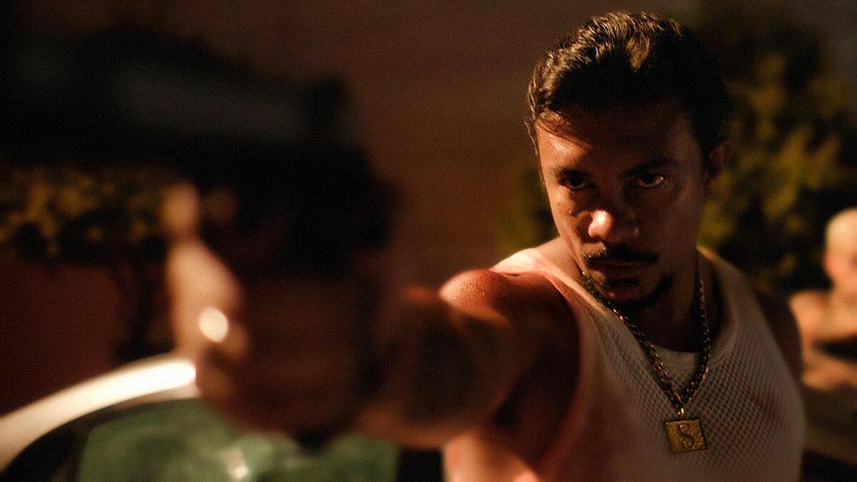 Xamã de camisa regata branca, segurando uma arma, apontando pra frente, durante gravação de filme