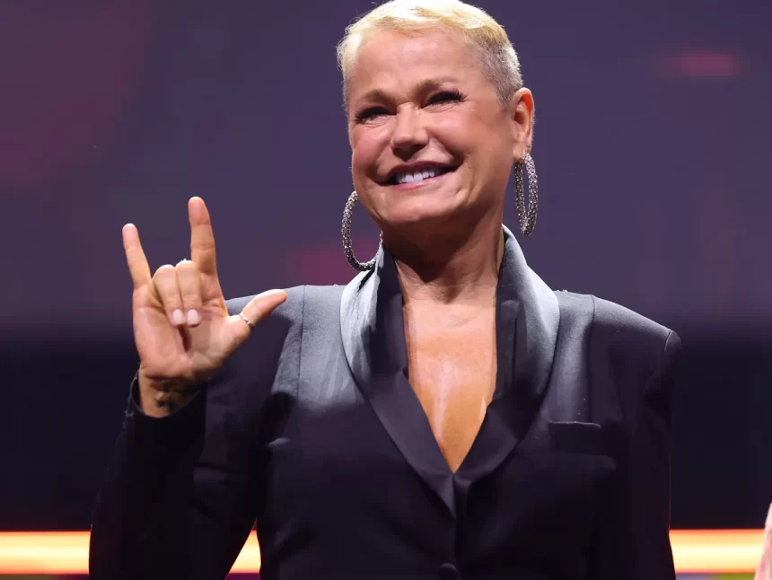 Xuxa de blazer preto, sorrindo, fazendo um simbolo com as mãos