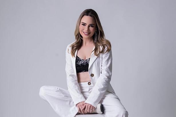 Rachel Sheherazade de calça e blazer branco com top preto sentada em um banco