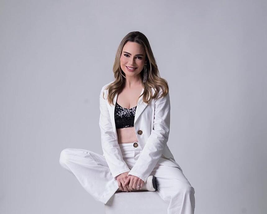 Rachel Sheherazade de calça e blazer branco com top preto sentada em um banco