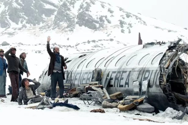 A imagem mostra parte de uma avião após queda em meio a neve com fuselagem aparente e jovens olhando para o céu pedindo socorro.